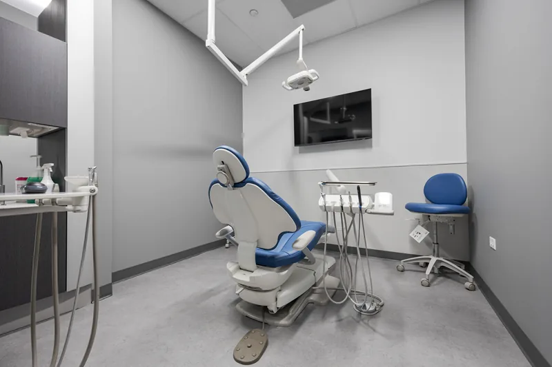 A treatment room with a blue dental chair, overhead light, and wall-mounted monitor
